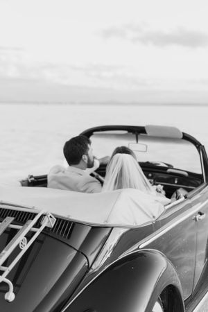 Black and white photo of a couple in a classic convertible car against a scenic backdrop.