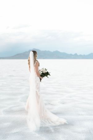 A bride in lace dress holds bouquet while standing on salt flats at sunset.