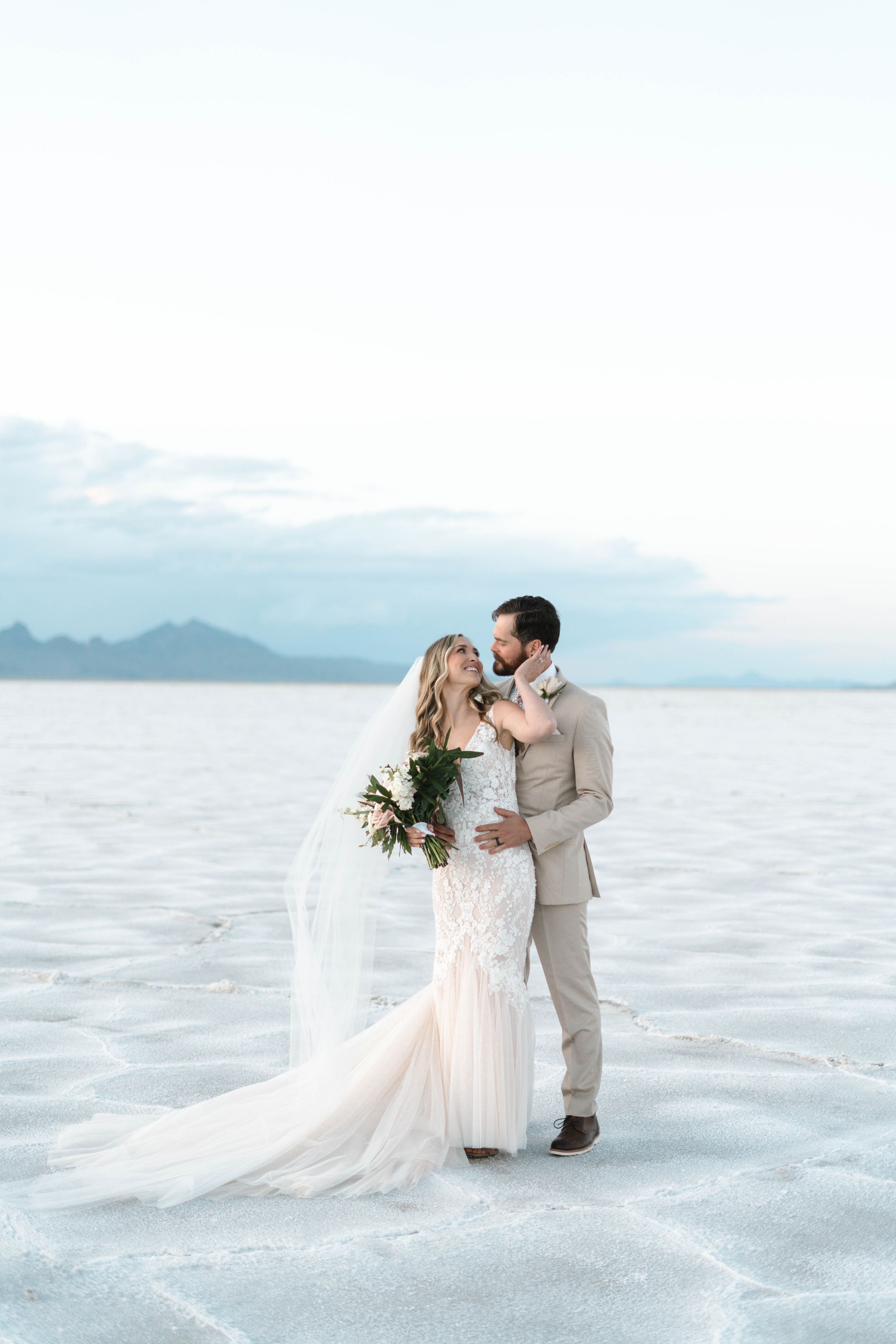 Wedding couple embraces on salt flats with mountain scenery during sunset.