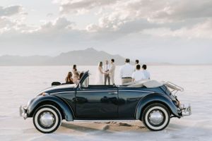 Vintage VW Beetle convertible parked on salt flats with wedding party in background.