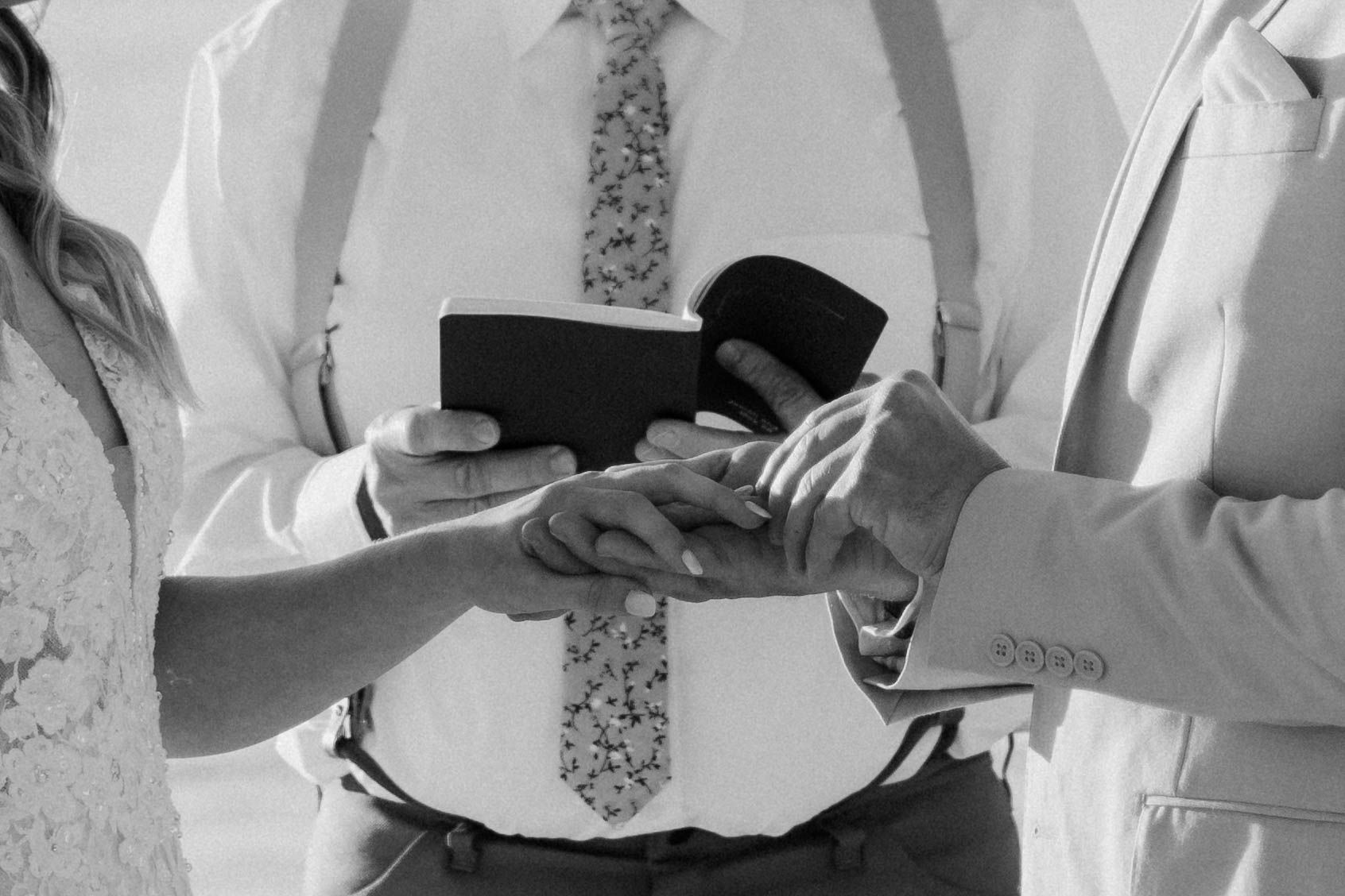 Black and white close-up of wedding ring ceremony with hands exchanging rings.