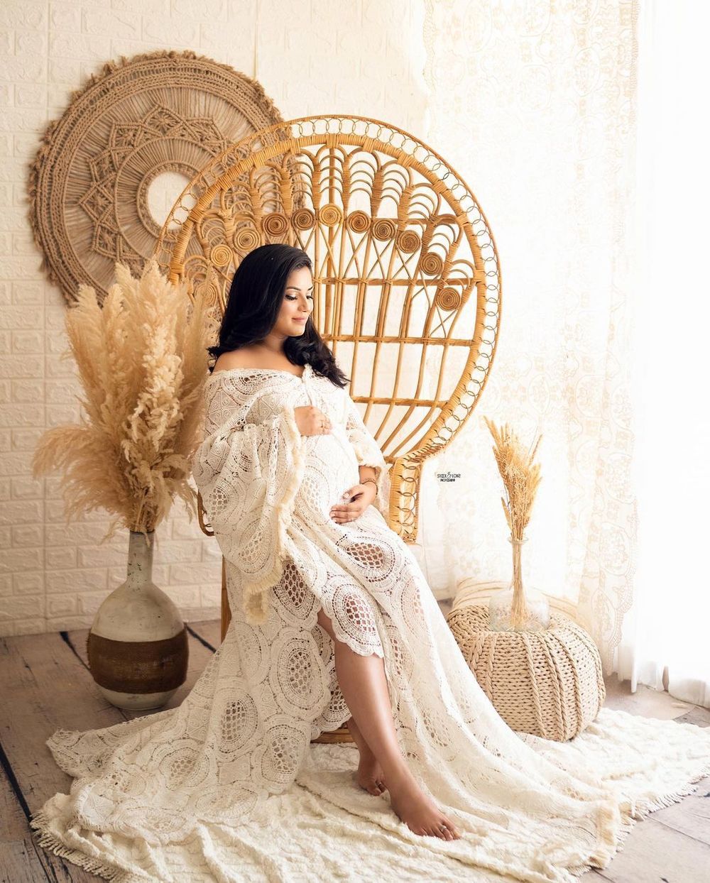 A maternity photoshoot featuring a white lace gown and boho decor with pampas grass and rattan furniture.