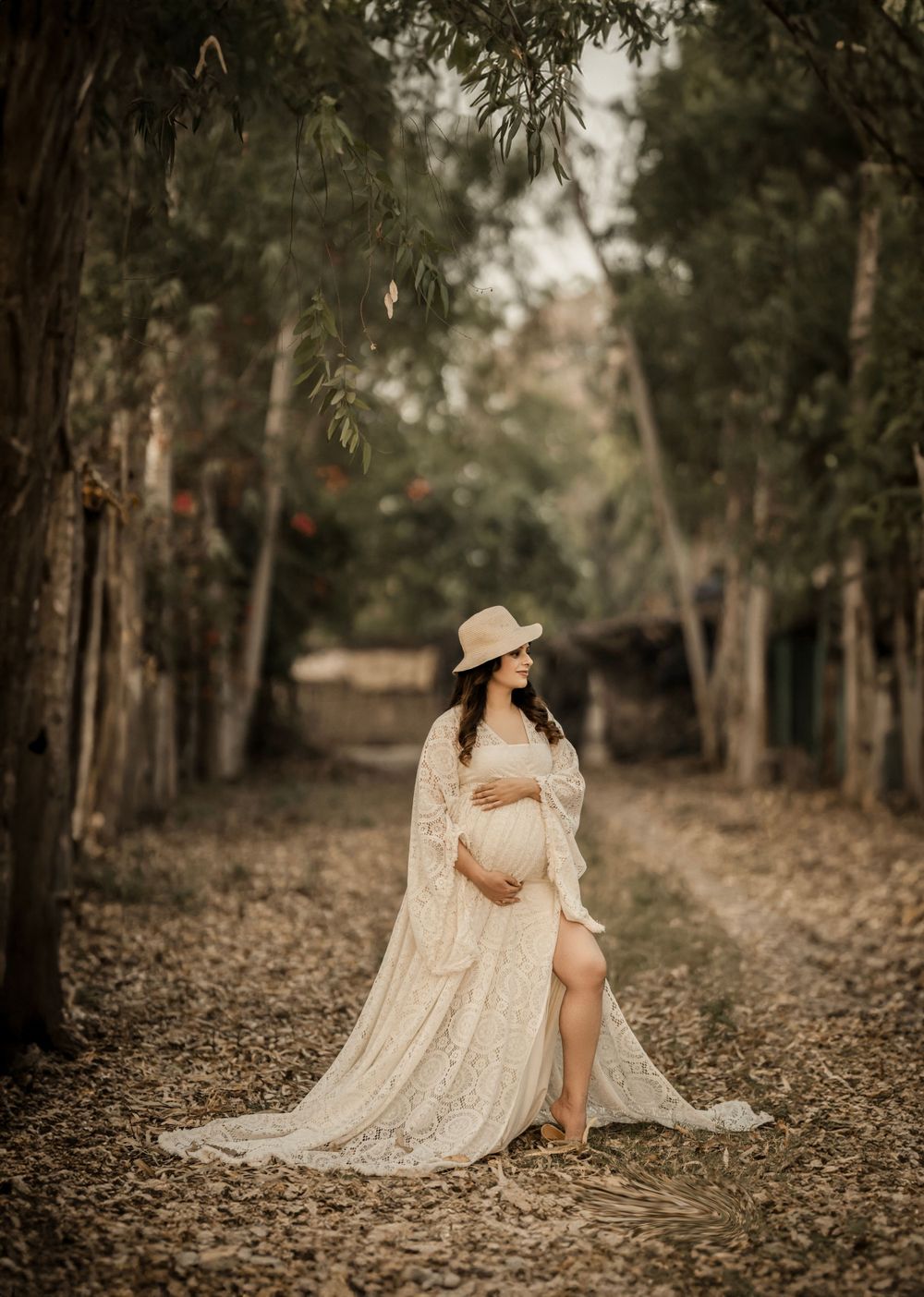 A maternity photo of an expectant mother in a lace dress and hat standing in a tree-lined path.