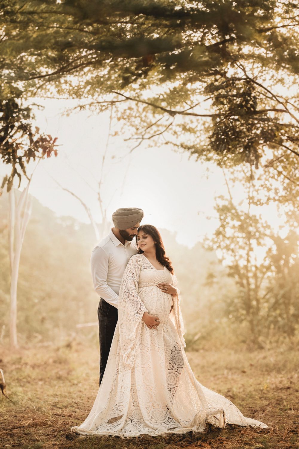 A romantic maternity photoshoot in a sunlit forest with soft golden light filtering through the trees.