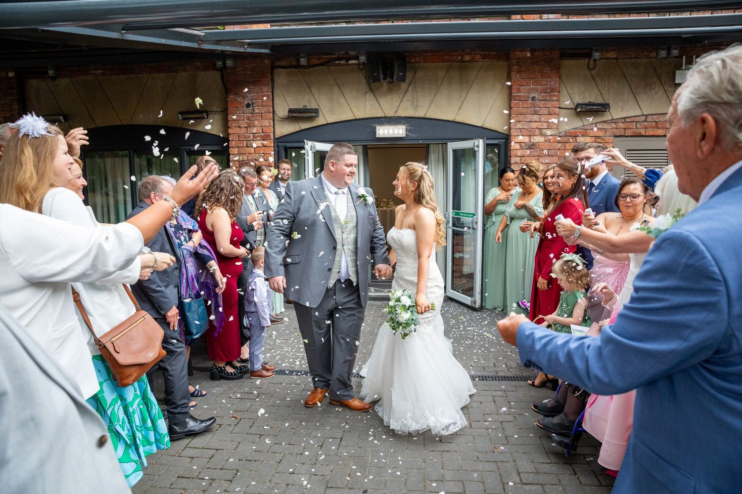 Bride and groom walk through confetti shower as wedding guests celebrate outside venue.