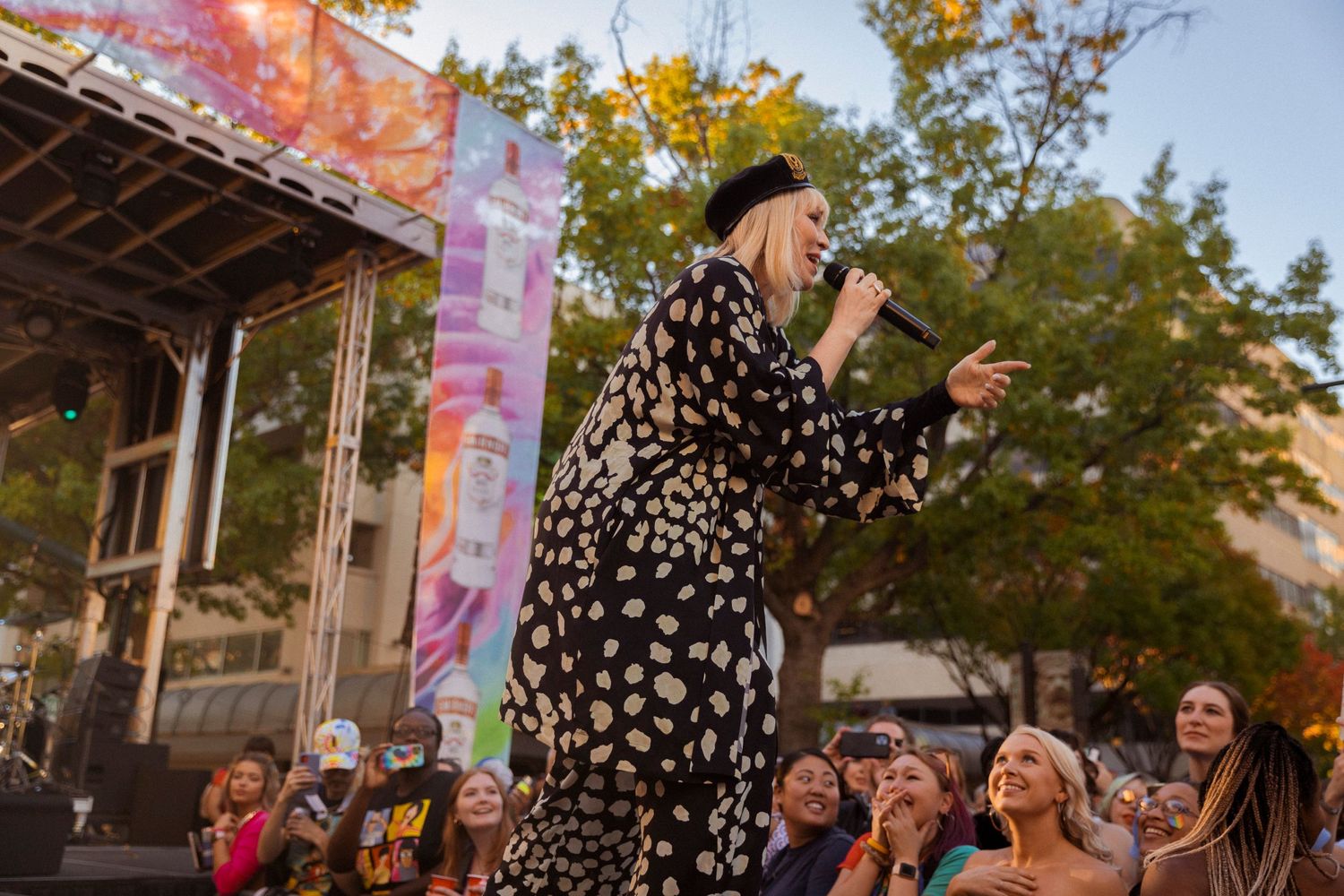 A performer in a polka dot dress sings on stage at an outdoor festival as the crowd watches in the evening light.