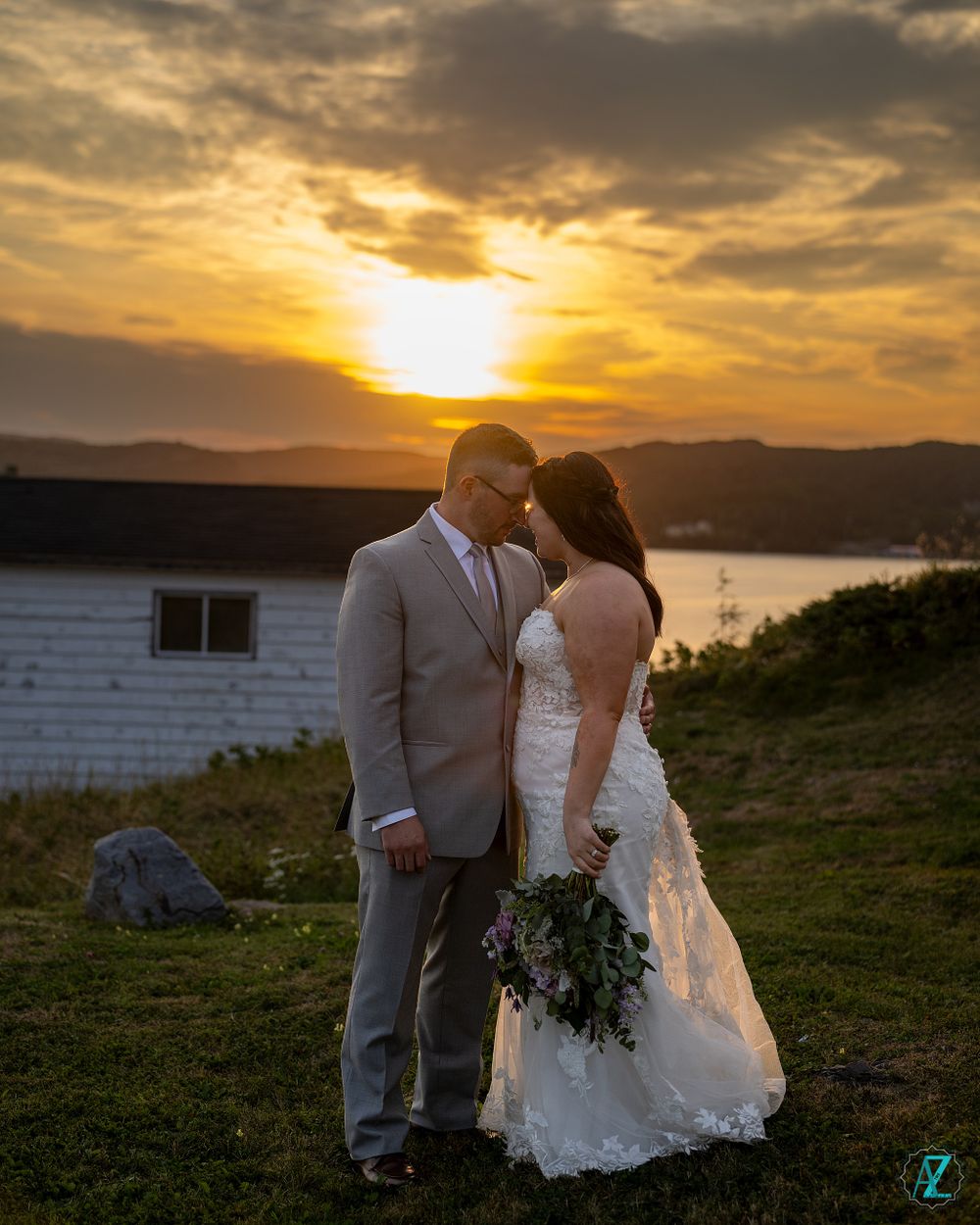 Couple holding bouquet poses during golden sunset near coastal building.
