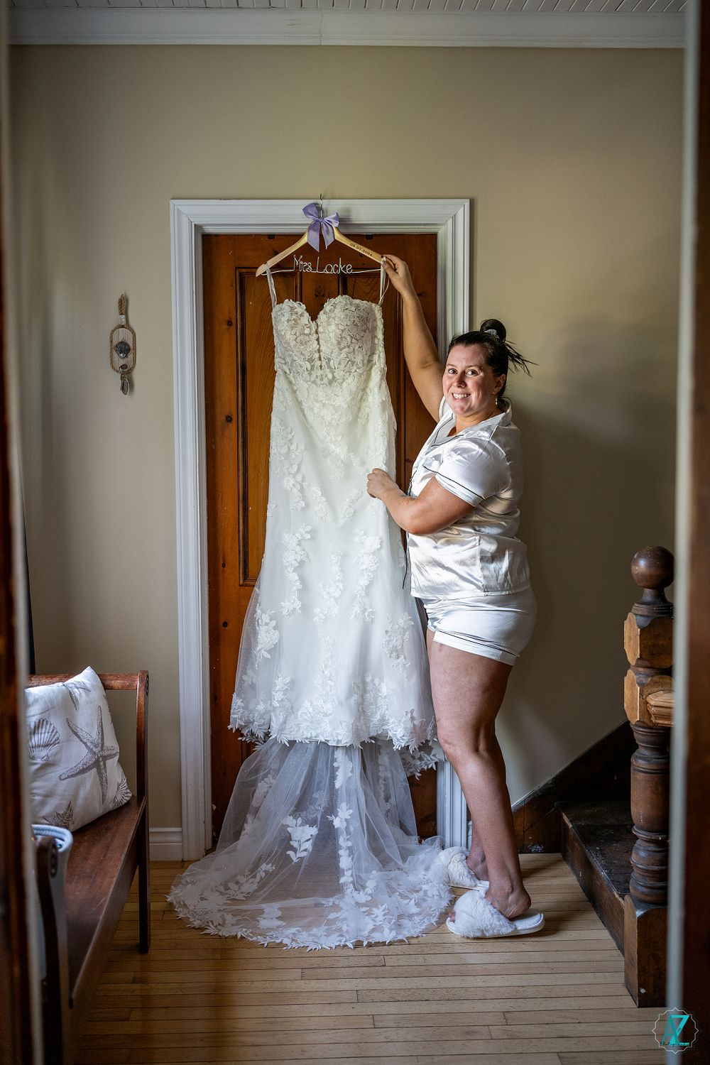 A detailed view of an elegant white lace wedding gown hanging on a wooden door in a hallway.