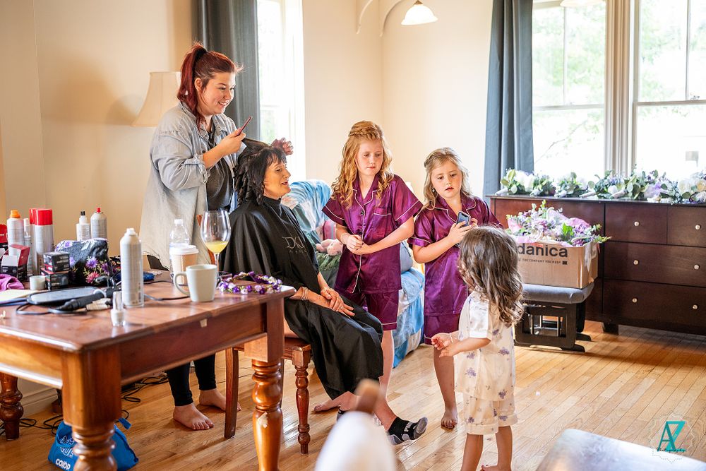 Multiple people coordinating hair and makeup preparations in a sunlit room with vintage furniture.