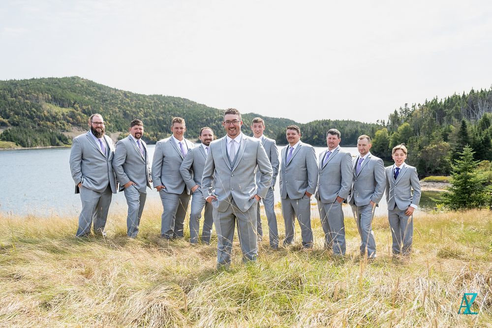 A wedding party in light gray suits stands in a line against a scenic lake and mountain backdrop.