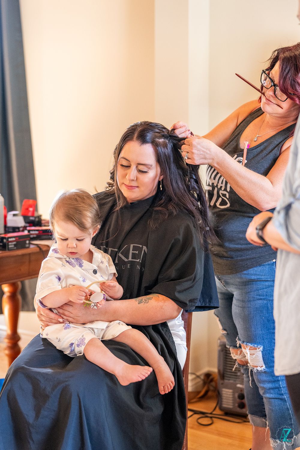 A wedding preparation scene shows a stylist working on someone's hair while holding a young child.