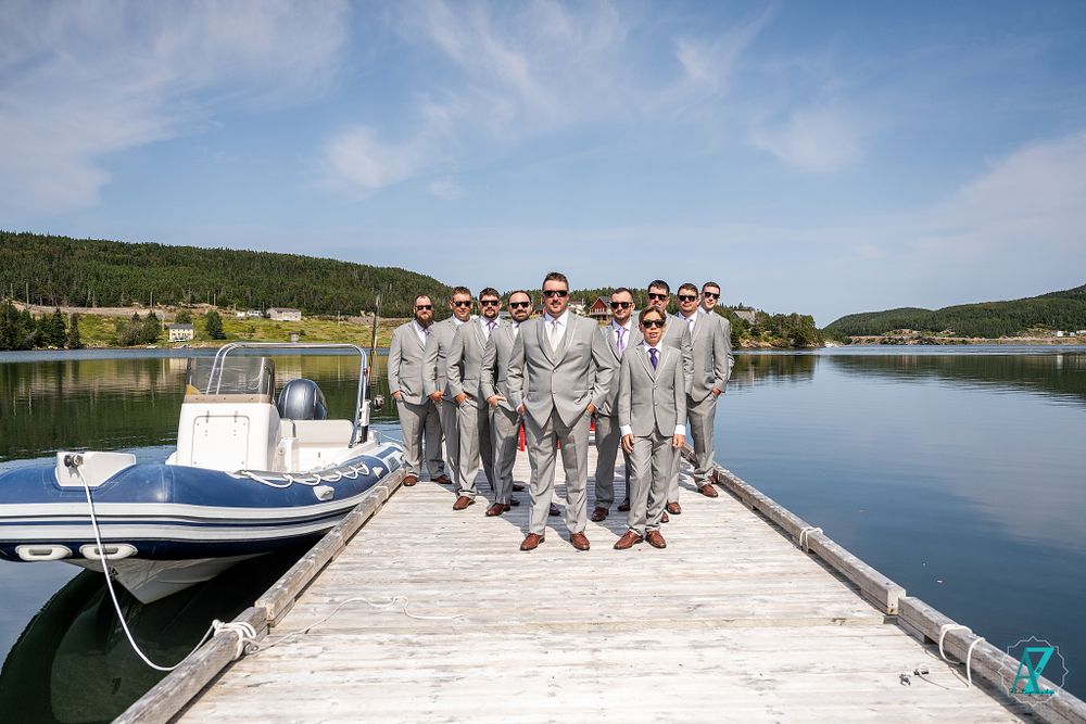 A wedding party in gray suits poses on a wooden dock with a boat and lake in the background.