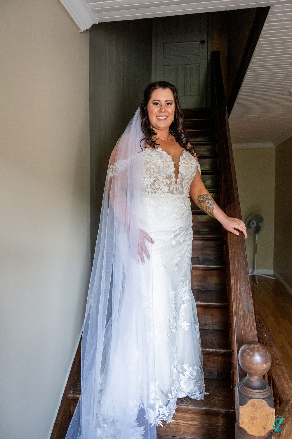 A bride poses on wooden stairs in a fitted lace wedding gown with long flowing veil.