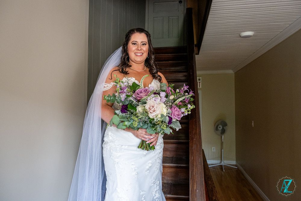 A bride in a white lace gown holds a pink and purple bouquet while standing on stairs.