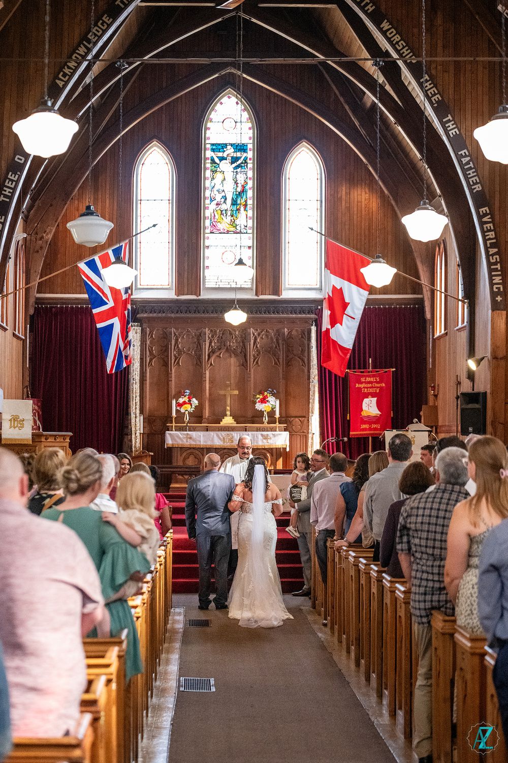 Church wedding ceremony with British and Canadian flags hanging beneath arched wooden ceiling.