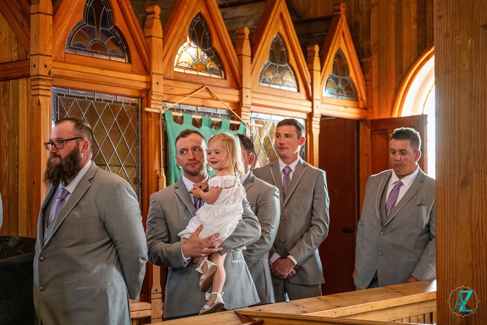 A wedding party stands in formal attire inside a church with gothic arched windows.