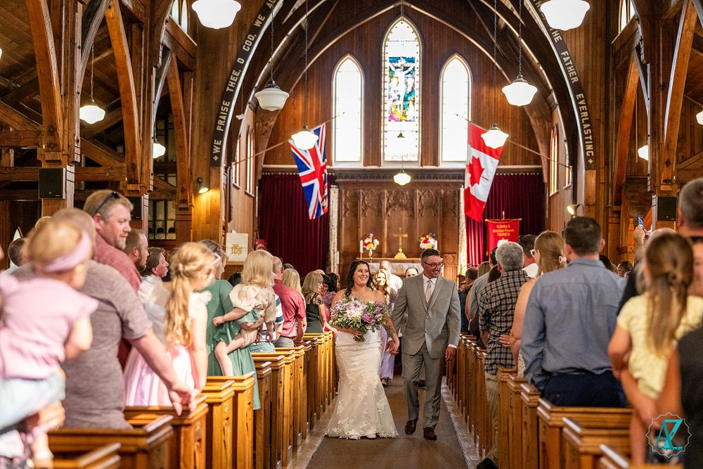Wedding guests stand in wooden pews as couple processes down church aisle under arched ceiling.