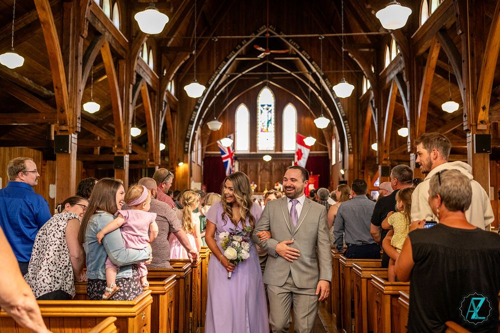 Wedding party walking down church aisle under vaulted wooden ceiling with glowing pendant lights.