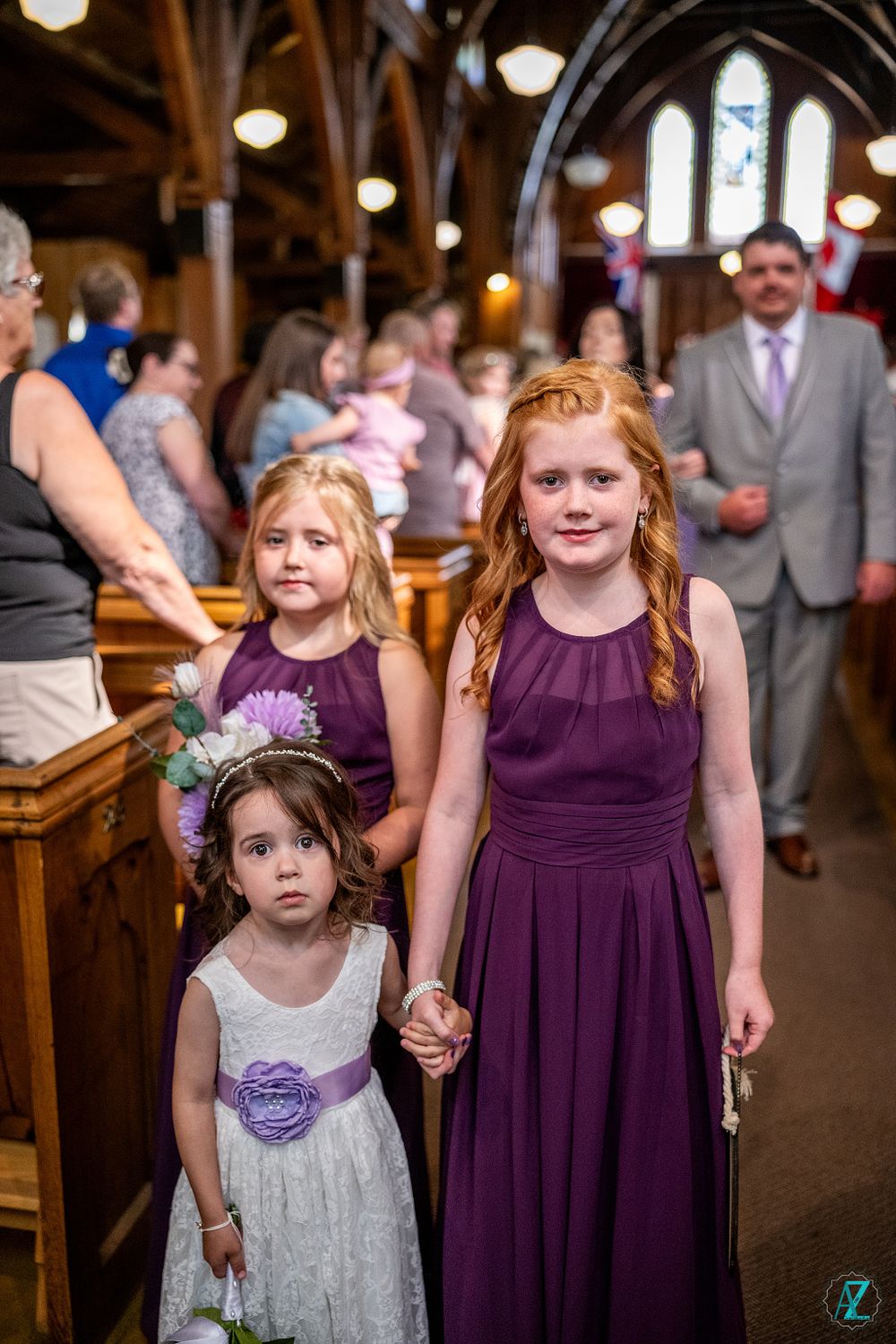 Young attendants in purple and white dresses stand in church aisle during wedding ceremony.
