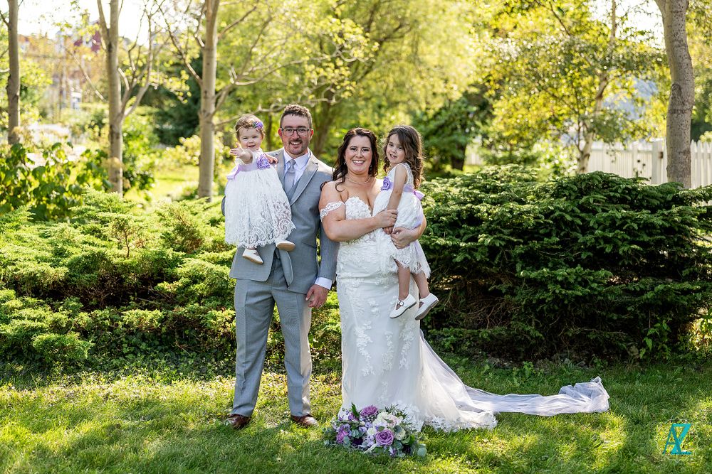 Wedding group standing in front of green hedges with purple flower bouquet on ground.