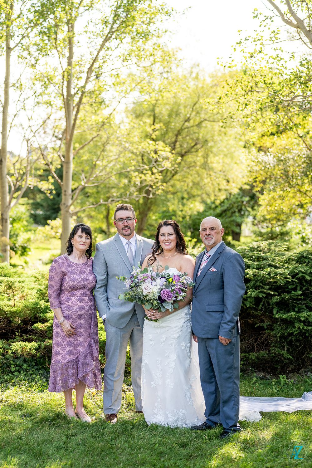 Wedding party poses in a sunny garden setting wearing a mix of purple and grey attire with floral bouquet.