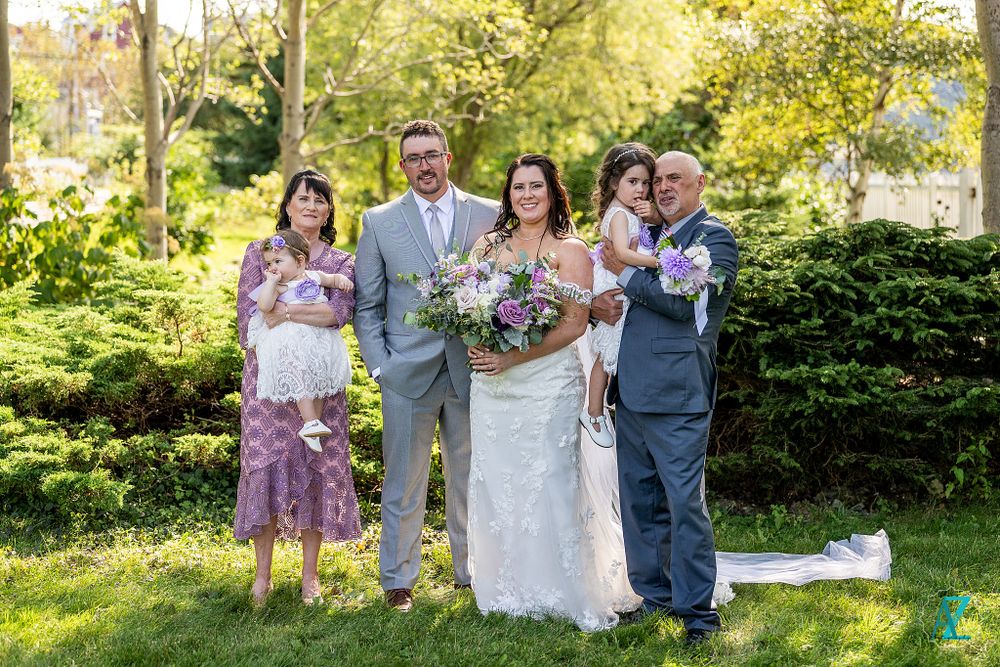 Formal wedding party portrait in garden setting with flowering shrubs and trees.