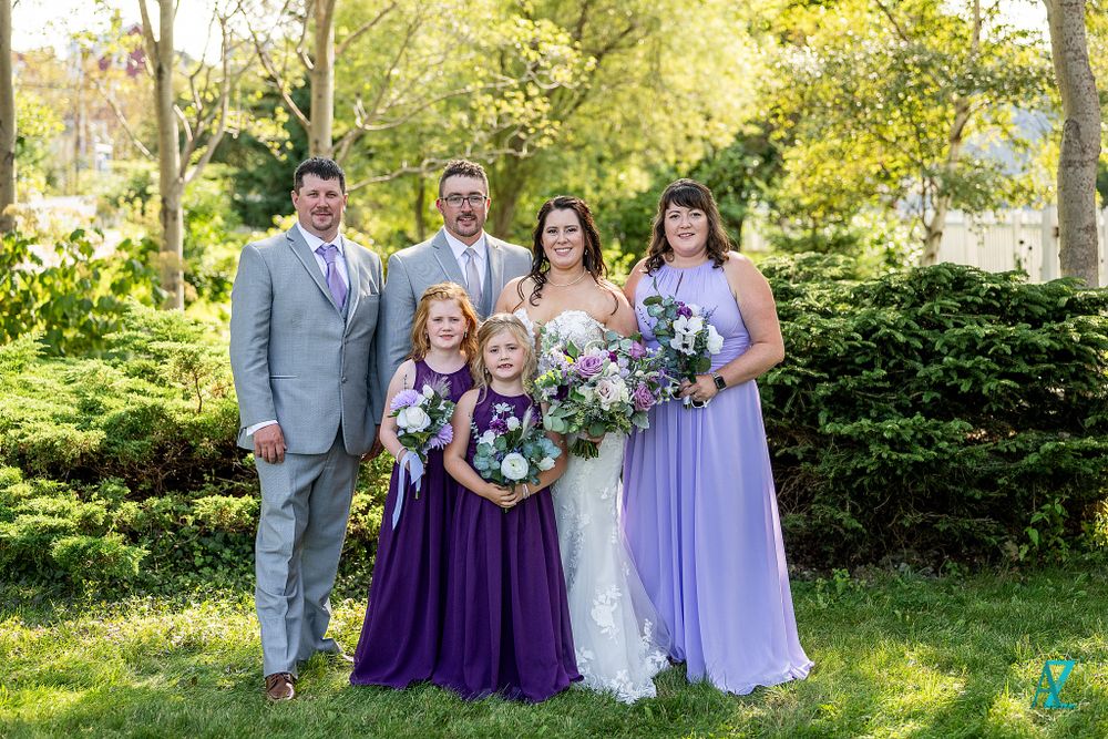 Wedding party poses in garden with matching grey suits and purple dresses holding floral bouquets.