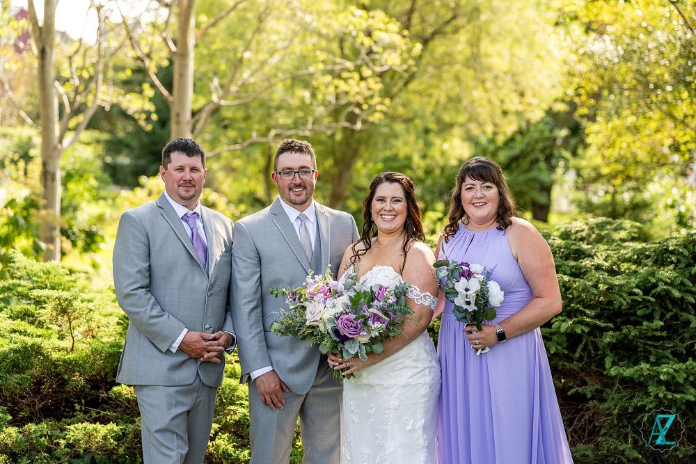 Wedding party members in grey suits and purple dresses pose against lush garden background.