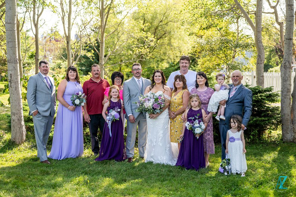 Large wedding group photo with mix of formal and casual attire in sunny garden setting.