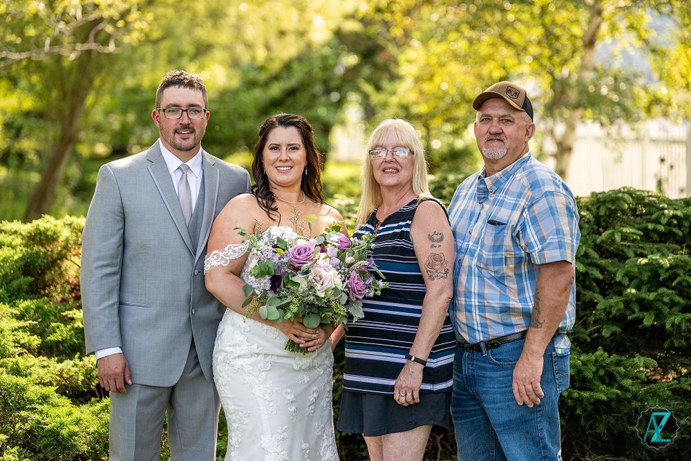 Wedding couple and family members gather for a portrait in front of evergreen shrubs.