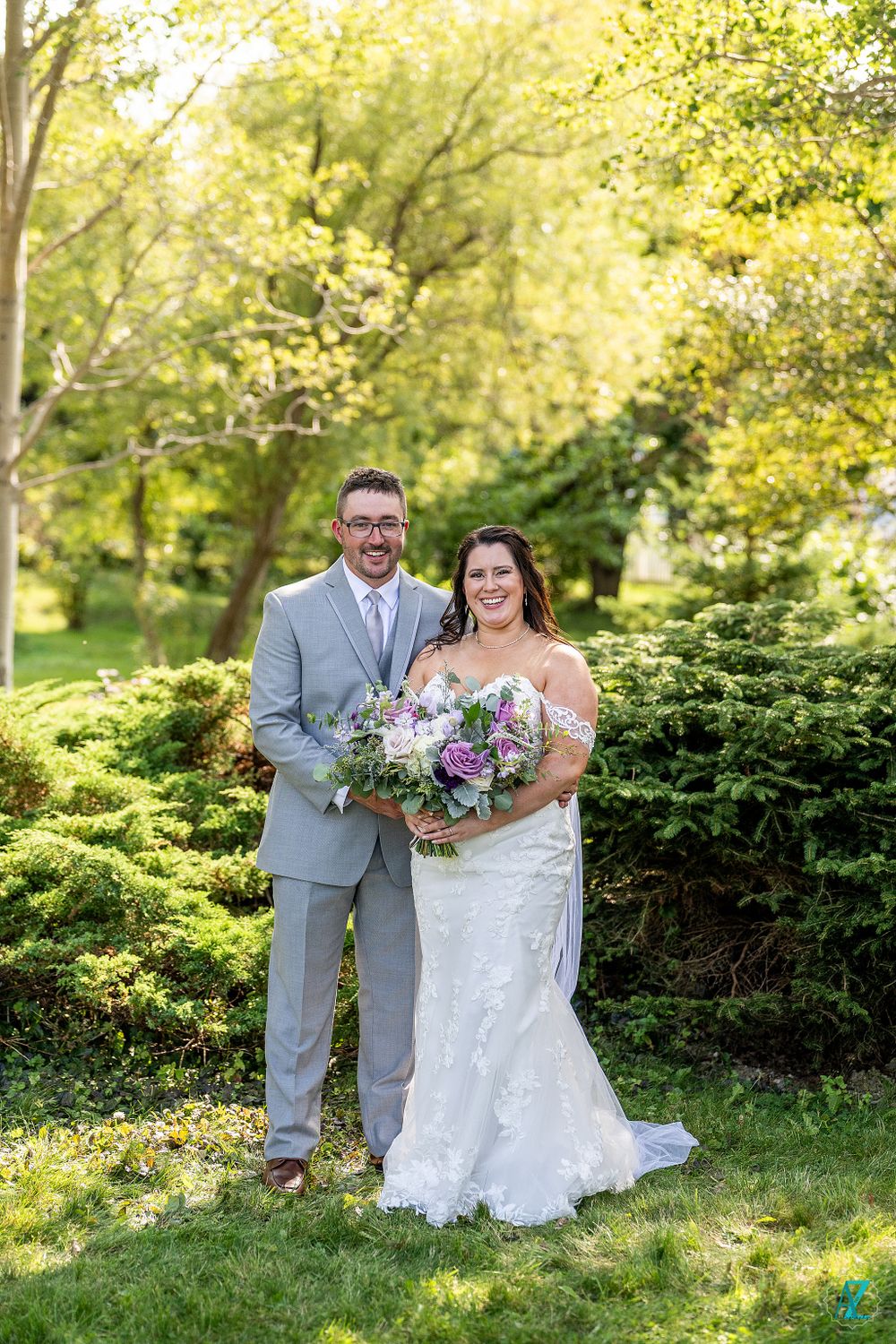 Wedding couple poses together holding purple and white flower bouquet in garden landscape.