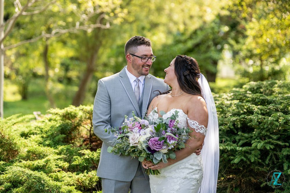 Newlywed couple shares intimate moment with purple bouquet in sunlit garden.