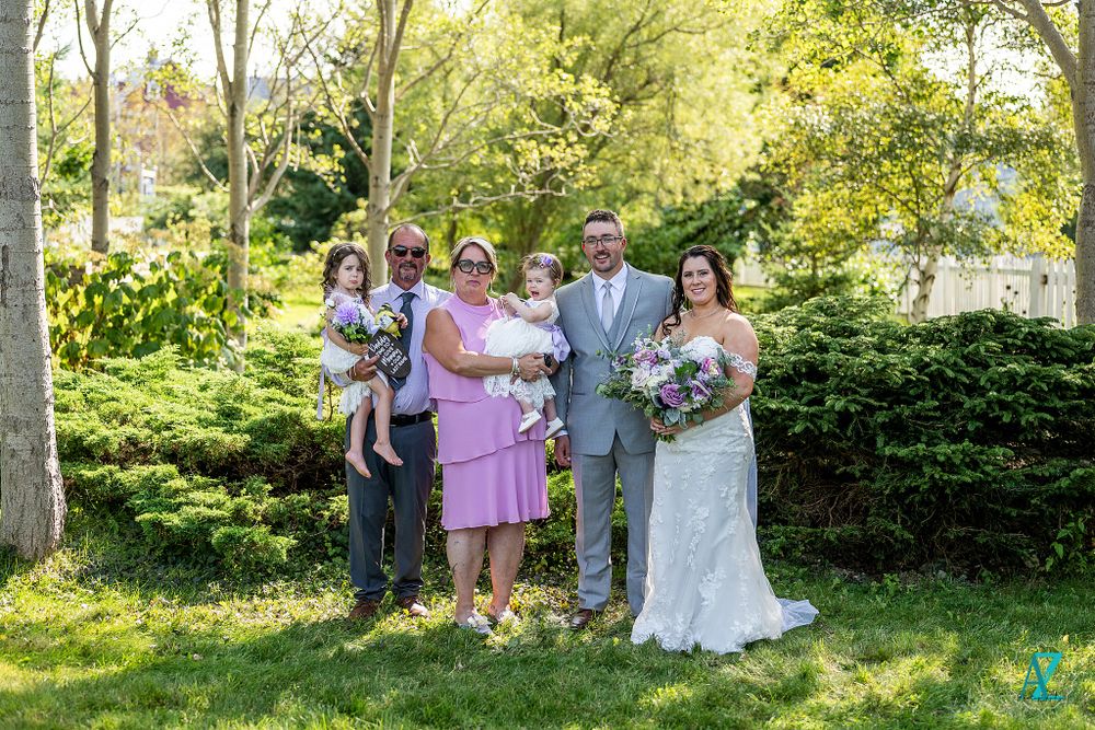Wedding party gathers for group photo in sunny garden with evergreen backdrop.