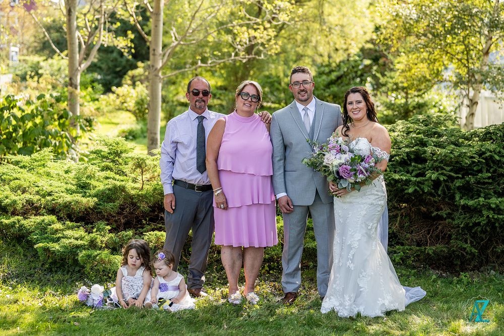 Wedding party poses with sitting children and floral bouquet in sunlit garden setting.