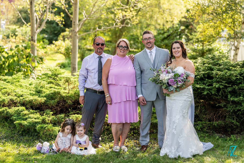 A wedding party poses outdoors with young children sitting on grass against a backdrop of lush green foliage.