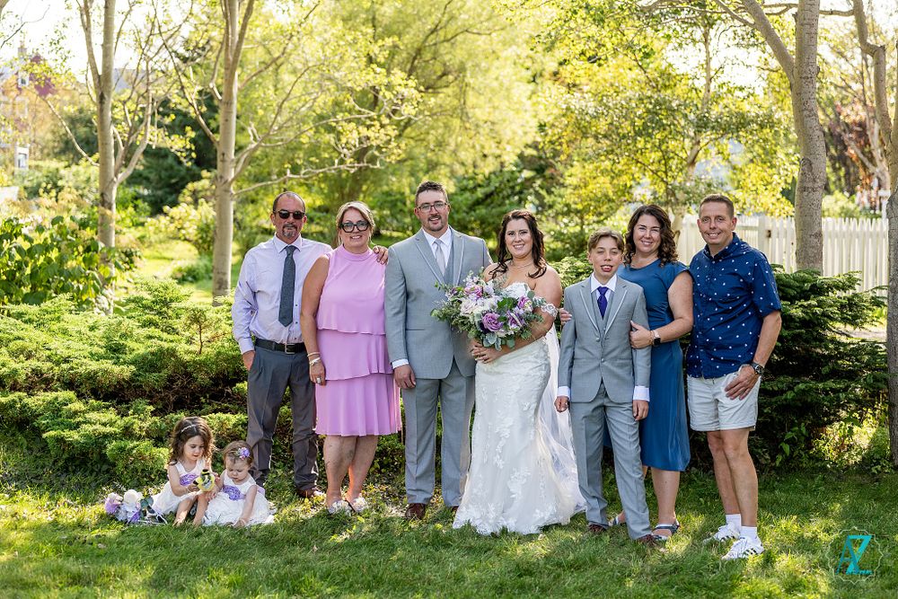 Extended wedding party stands together for a group photo in a sunny garden setting.