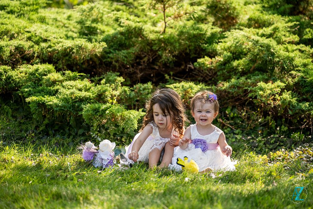 Two children in white dresses play and laugh on grass during a sunny afternoon.