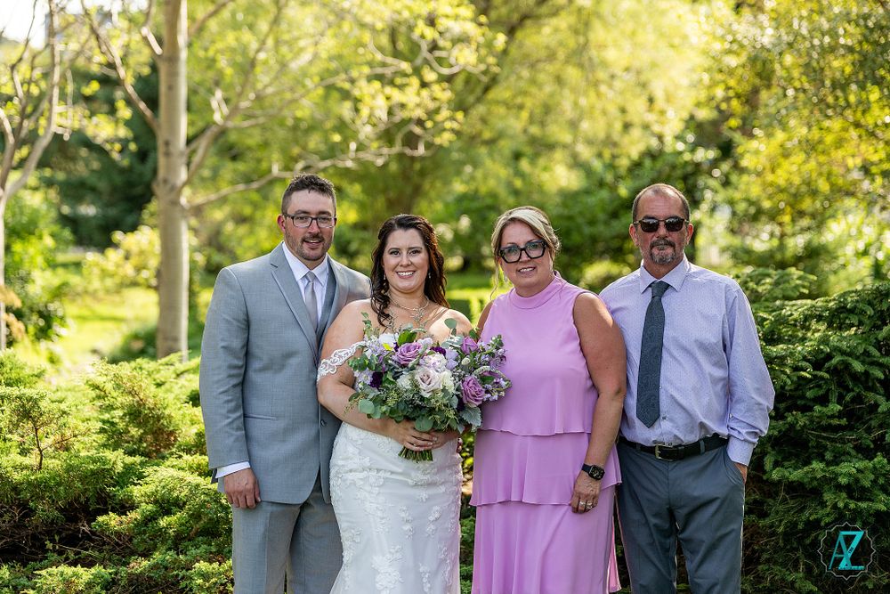 Small wedding party group poses together with bride holding purple and white bouquet in garden setting.