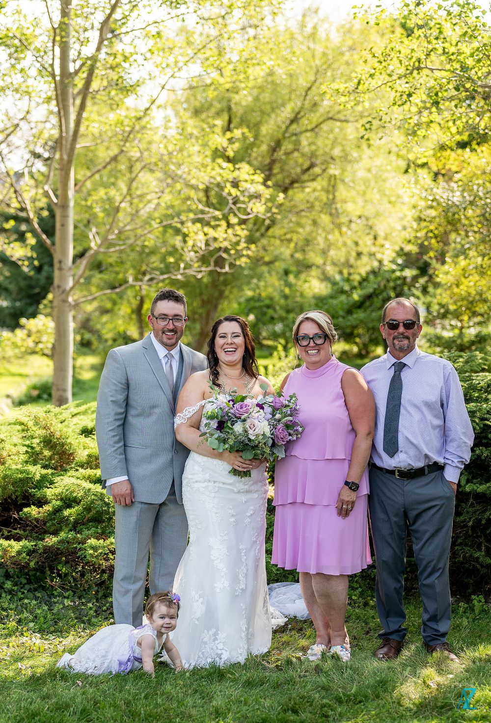Small wedding group poses outdoors with child sitting on ground in front.