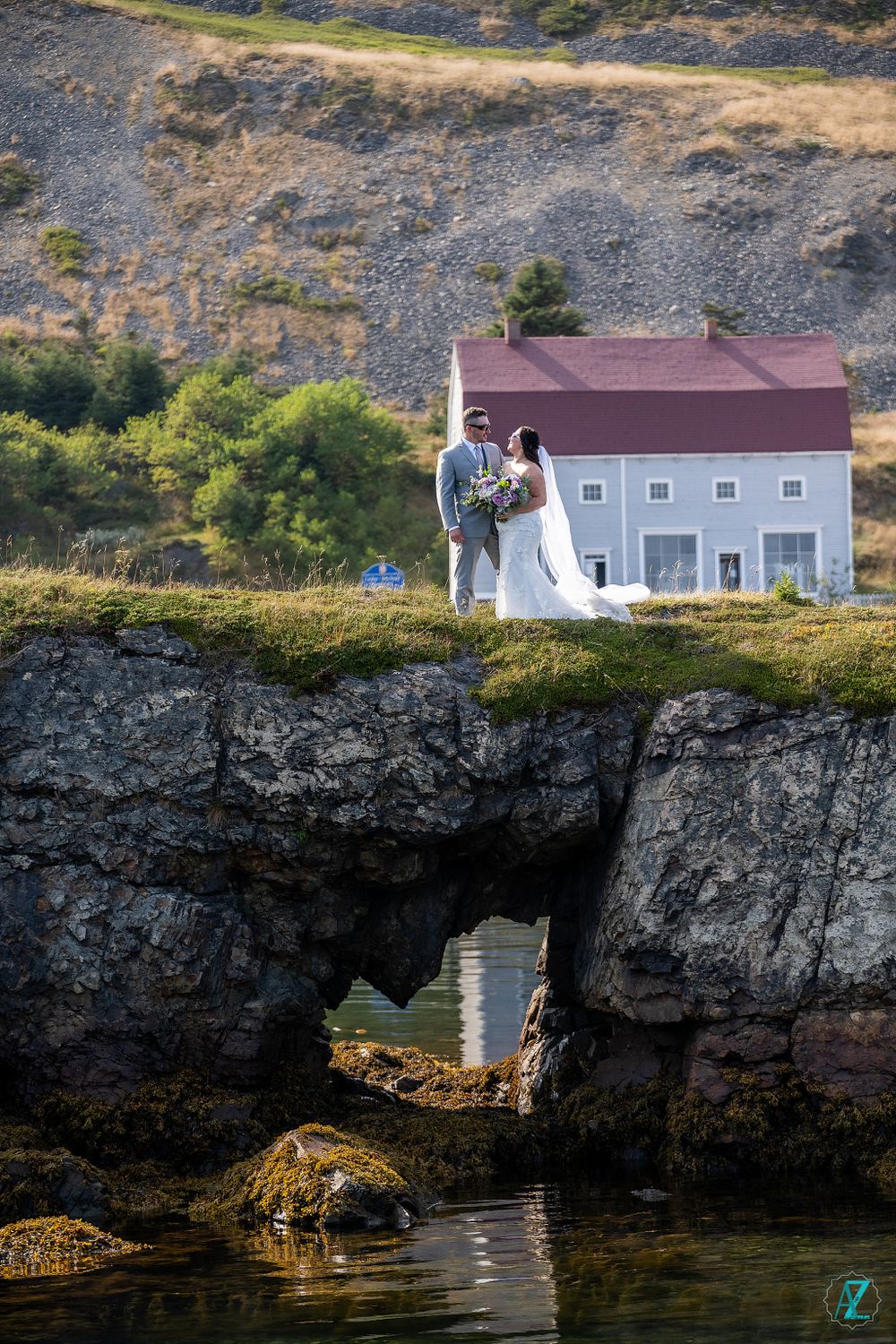 Wedding couple poses on dramatic cliff edge with white house and ocean in background.