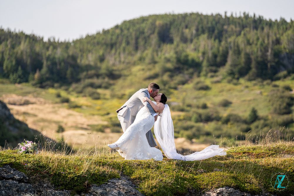 Wedding couple embraces on windswept coastal cliff with flowing dress and mountain backdrop.