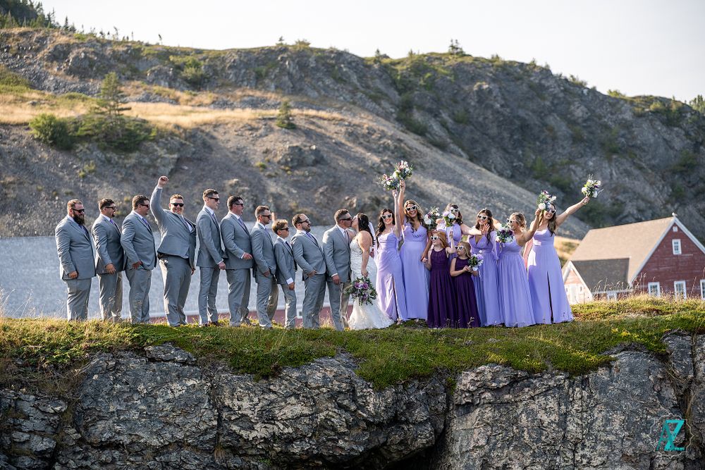 Wedding party in purple and gray attire poses dramatically on coastal cliff edge.