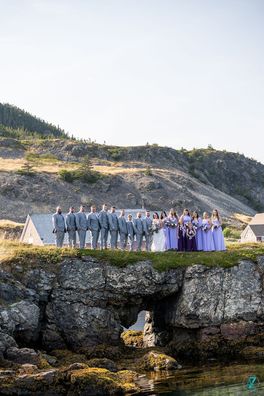 Large wedding party stands on dramatic rocky cliff overlooking ocean inlet and coastal houses.