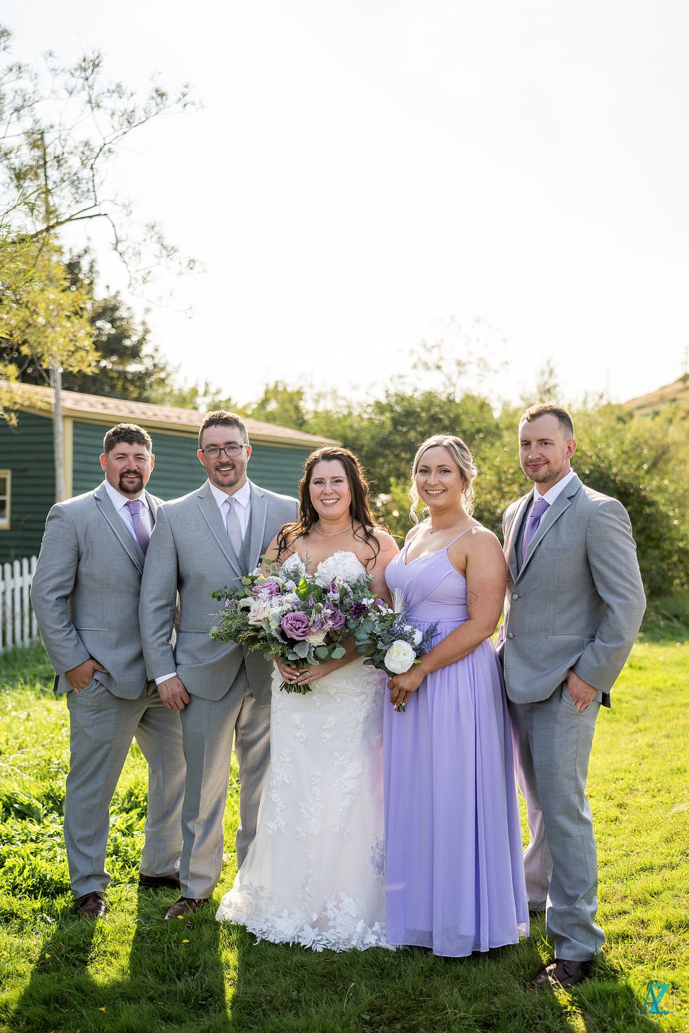 Wedding group in gray suits and lavender dresses poses against green building backdrop.