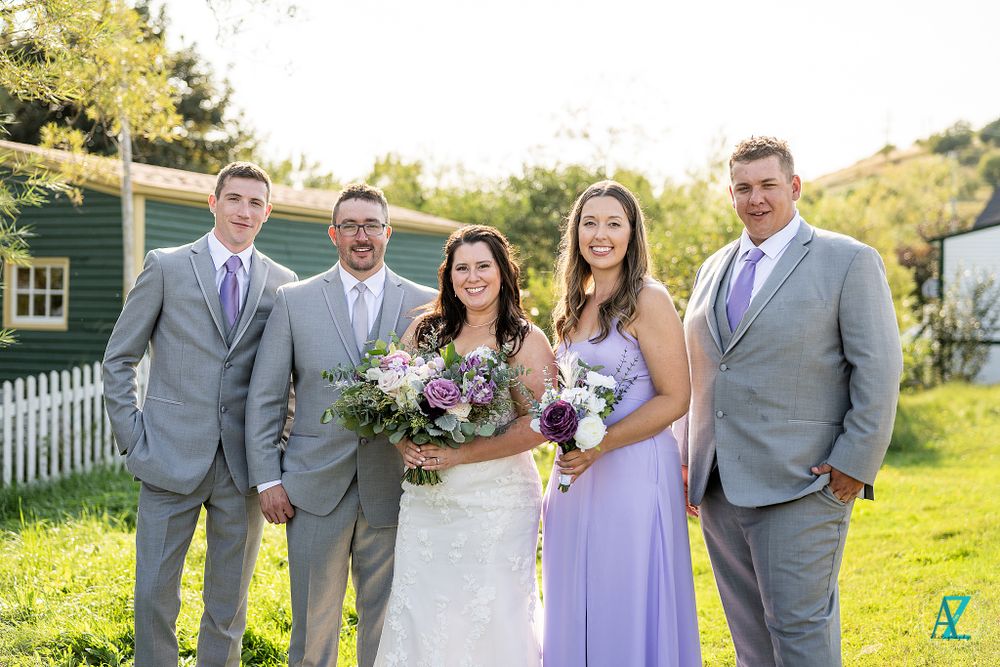 Wedding party in coordinated gray suits and lavender dresses poses on green lawn.