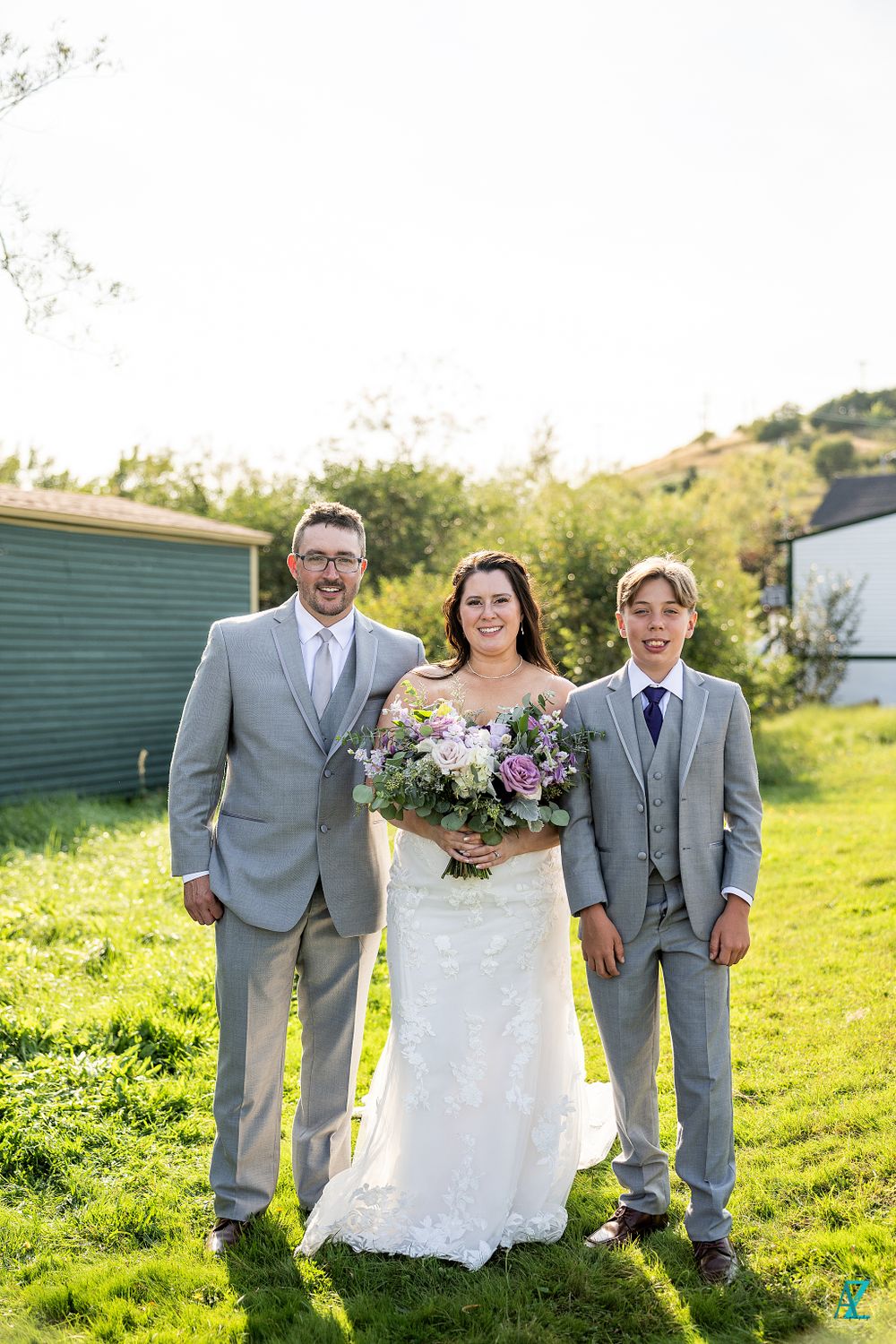 Wedding photo with three people in formal attire posing on green lawn with building in background.