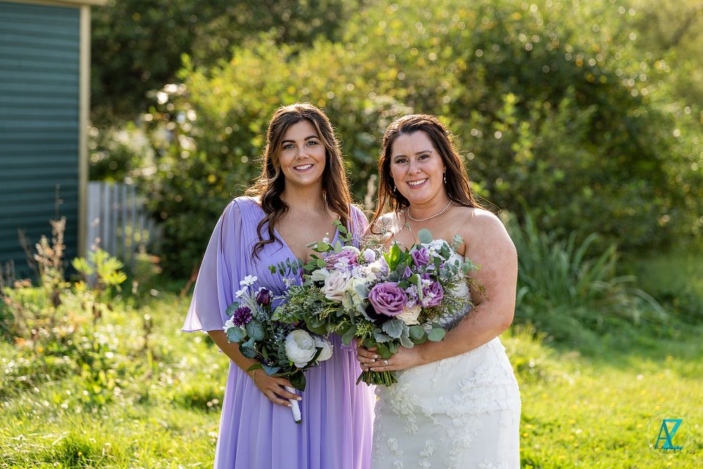 Two wedding attendants holding purple and white flower bouquets pose in a sunny garden setting.