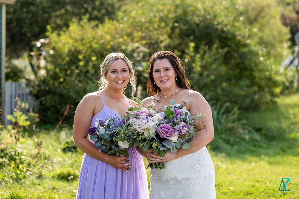 Two wedding attendants pose with matching purple and white bouquets against sunny garden backdrop.