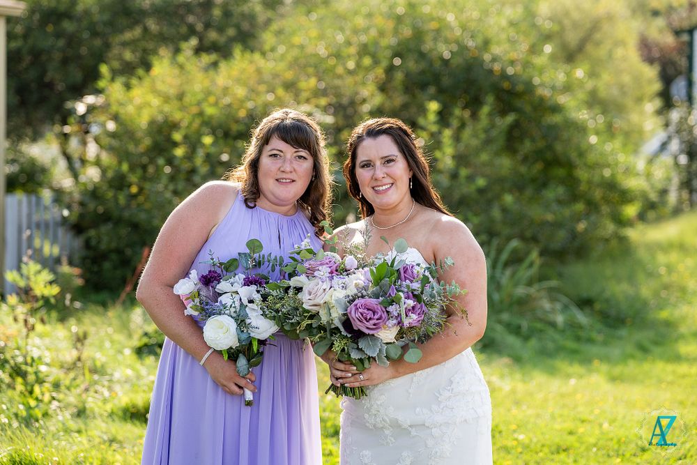 Two wedding party members smile while holding coordinating purple and white floral arrangements outdoors.