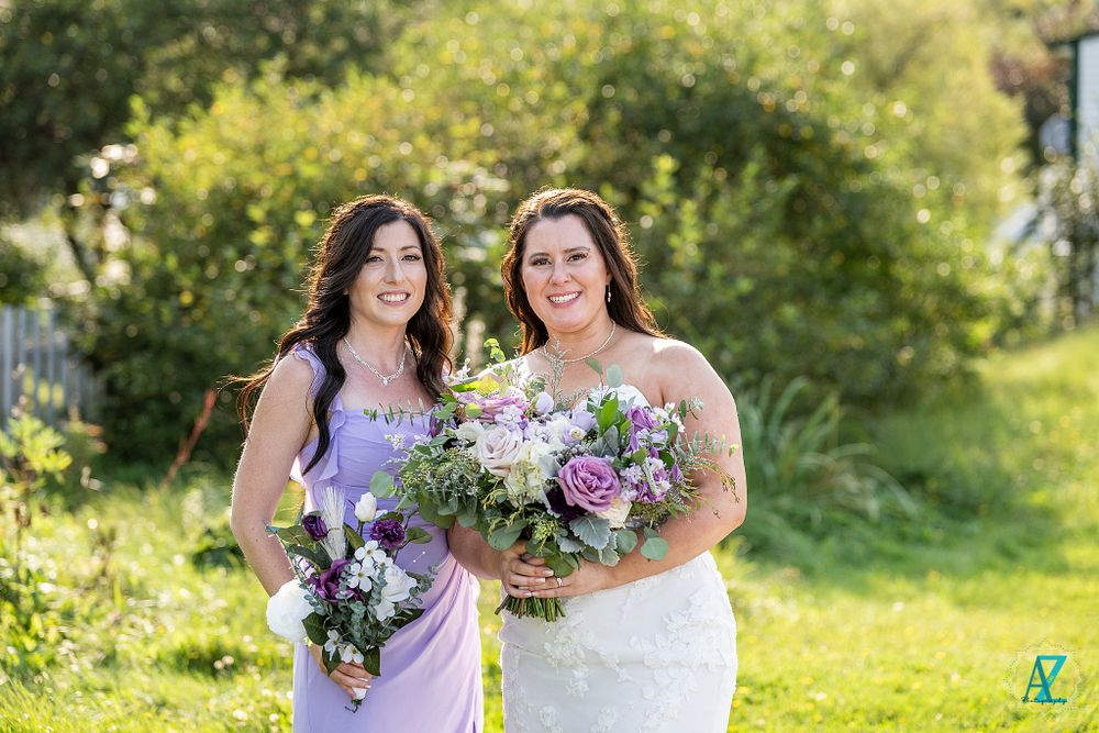 Wedding attendants in white gown and lavender dress pose with matching bouquets in garden setting.